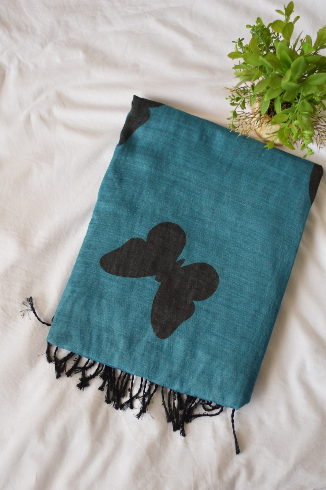 Teal Chanderi Linen Cotton saree with black butterfly design on a white surface with a plant in the corner.