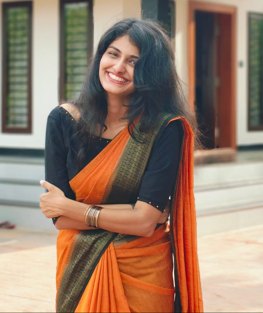 Woman in an orange saree standing in front of a traditional house