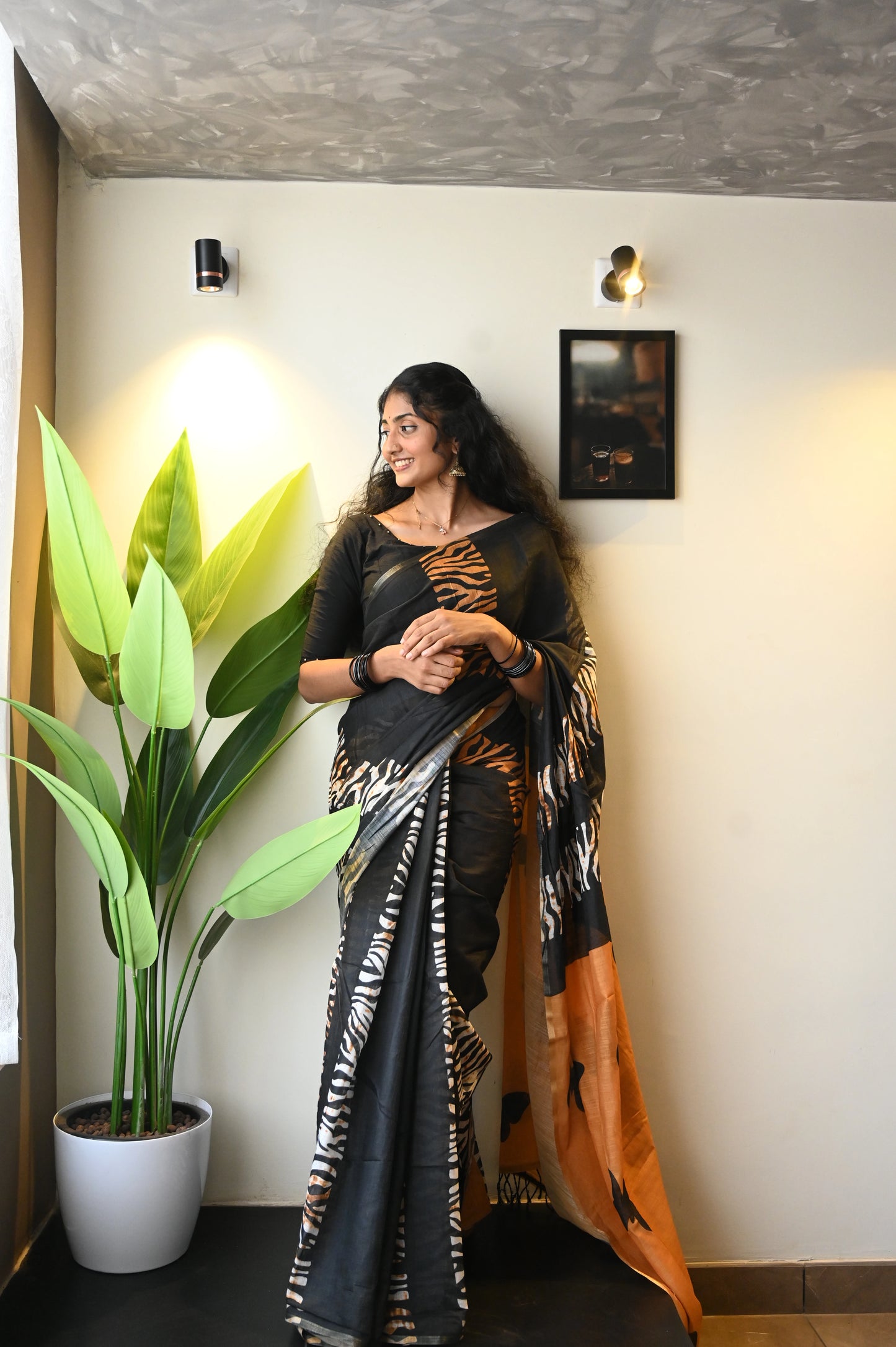Women wearing Shalabha_Tiger Stripe & Butterfly Patchwork Saree in a black and orange combo from saree  from thambai standing indoors with a plant and wall decor.