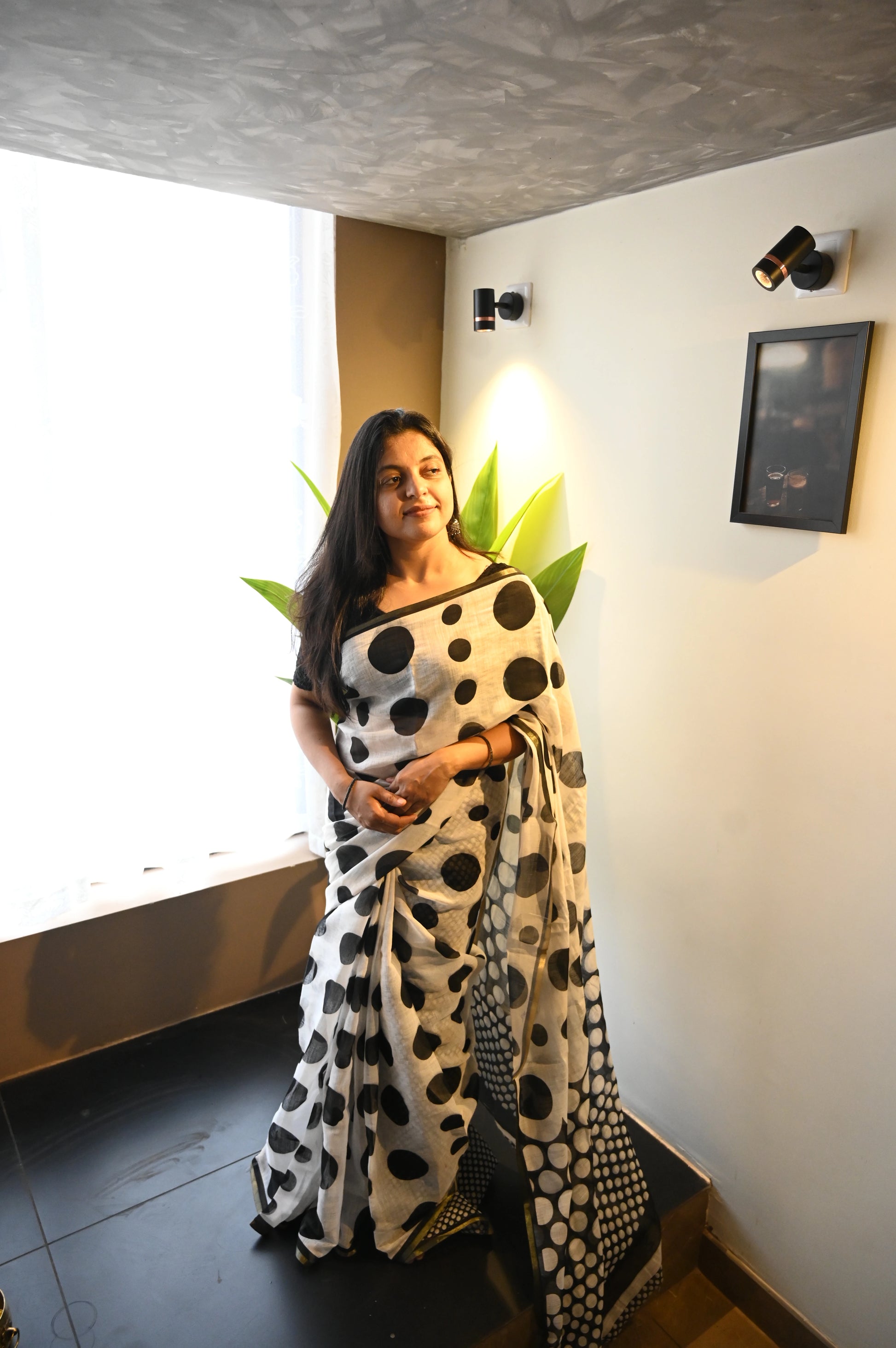 women wearing saree from thambai's Ambili_Polka dot print semi-linen cotton saree in a dark green  and white patterned combo standing indoors near a window.