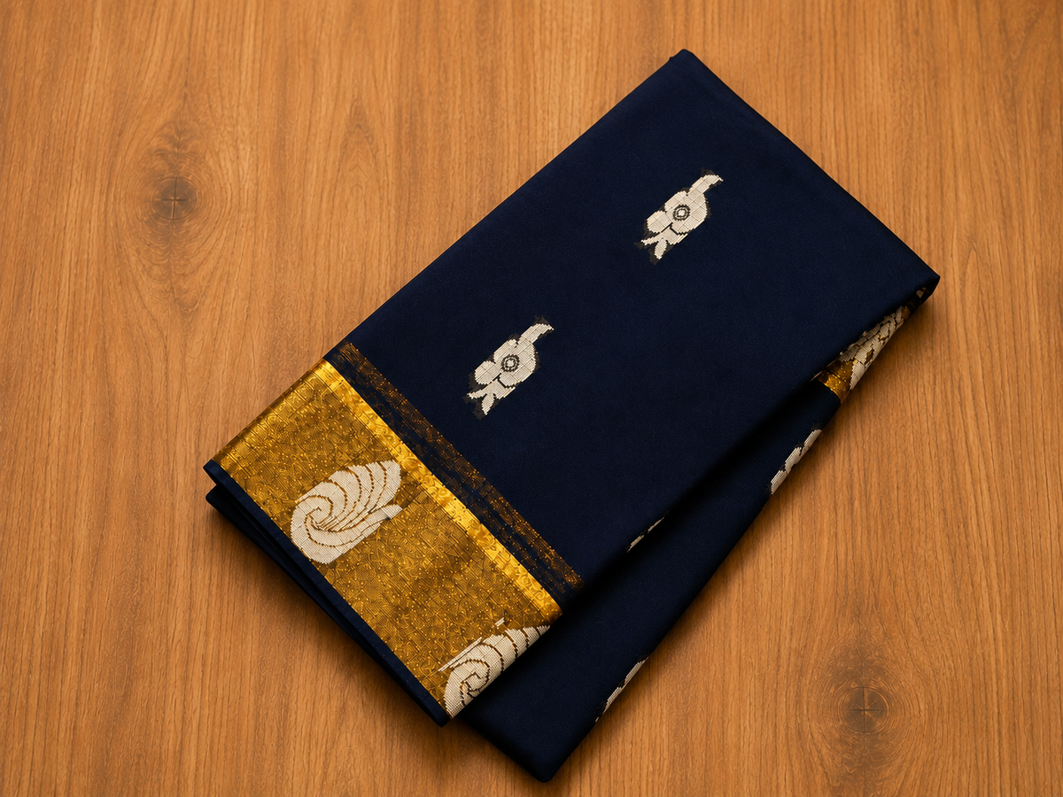Neatly folded deep navy blue saree featuring a rich gold woven zari border and subtle silver silver-white motifs, displayed flat against a smooth wooden background from Saree from Thambai online saree store.