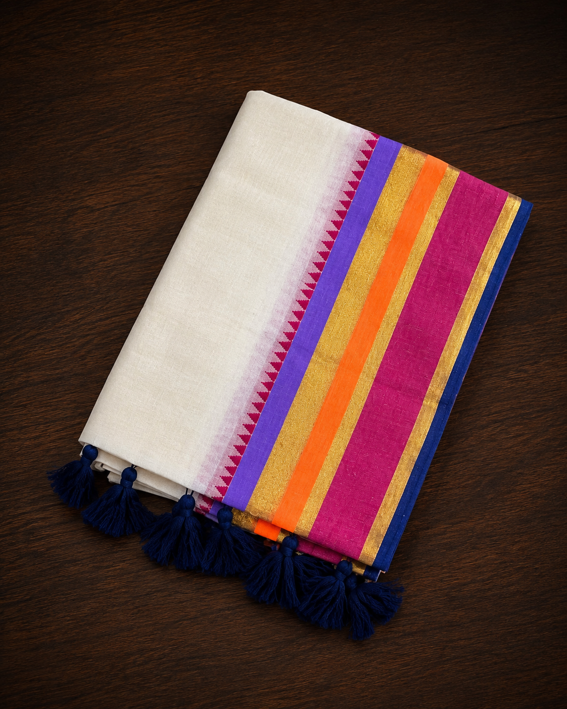 A folded off-white mul cotton saree featuring a vibrant, multi-colored border with parallel stripes of lavender-purple, shimmering gold, bright neon orange, and hot pink, accented by a tiny pink temple design edge and finished with thick navy blue tassels on a warm wooden tabletop.