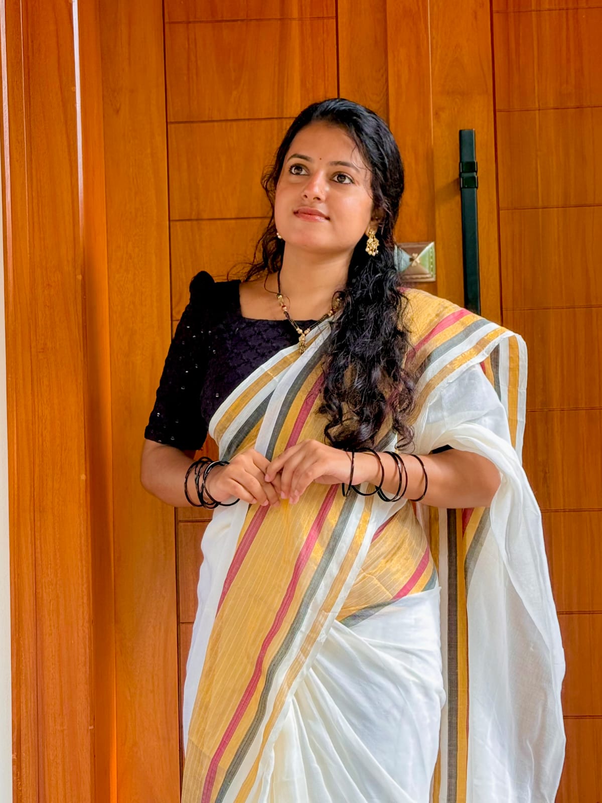 A woman modeling the Thambai Onam Special Malar Kerala Saree, featuring a traditional off-white cotton body with an elegant gold, black, and maroon striped border.