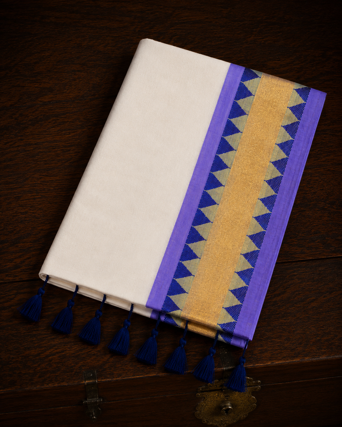 A folded off-white mul cotton saree showcasing a lavender-purple border with a central metallic gold kasavu band, accented by rows of deep blue geometric triangle temple patterns, and finished with thick royal blue tassels on a polished wooden surface.