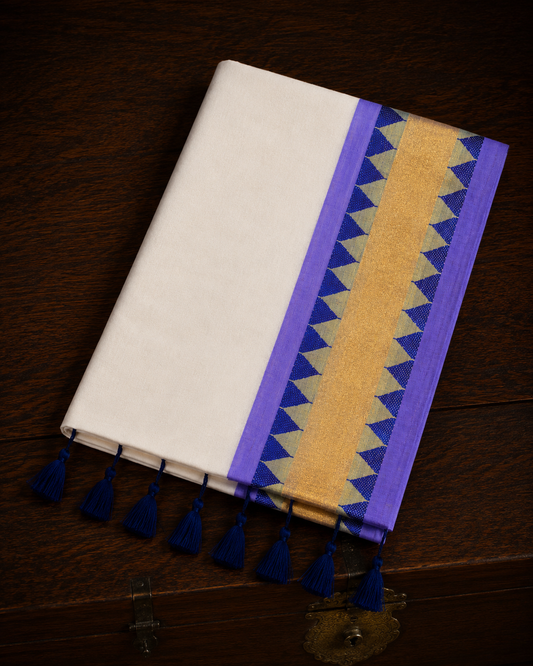 A folded off-white mul cotton saree showcasing a lavender-purple border with a central metallic gold kasavu band, accented by rows of deep blue geometric triangle temple patterns, and finished with thick royal blue tassels on a polished wooden surface.