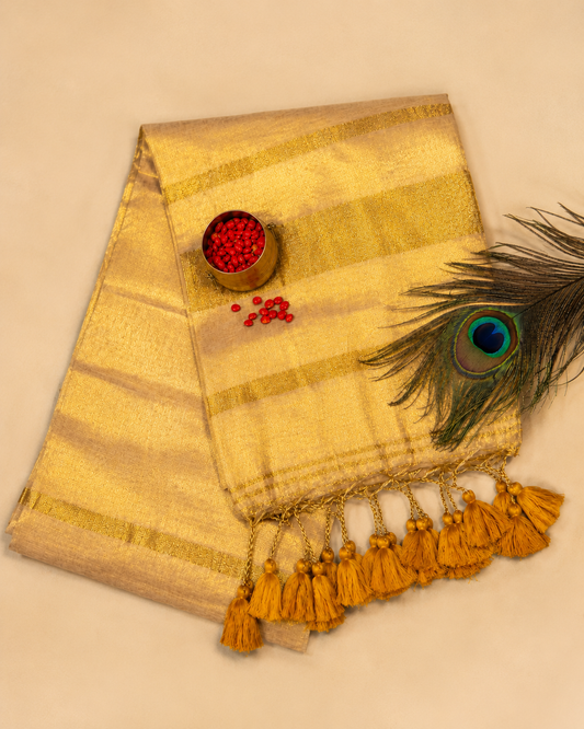 A luxurious, metallic gold mulcotton onam Kerala saree with self-textured horizontal bands and heavy mustard-gold tassels, styled beautifully on a beige background alongside a peacock feather, a small brass bowl filled with bright red manjadikuru seeds