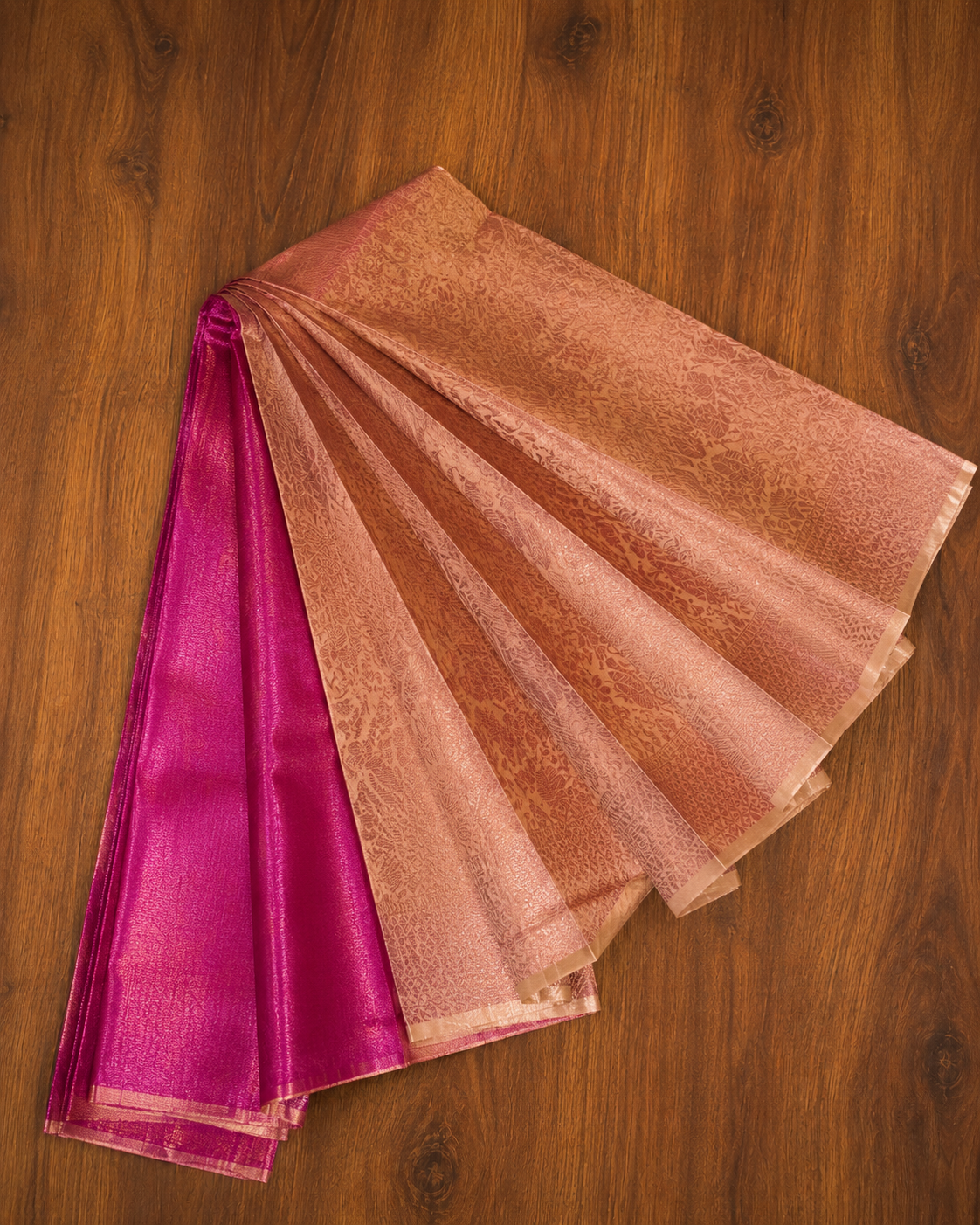 A folded and fanned-out kubera patt saree or fabric piece displayed on a dark wood background, showing a warm peach-beige body with subtle zari embroidery and a contrasting bright magenta-pink border.