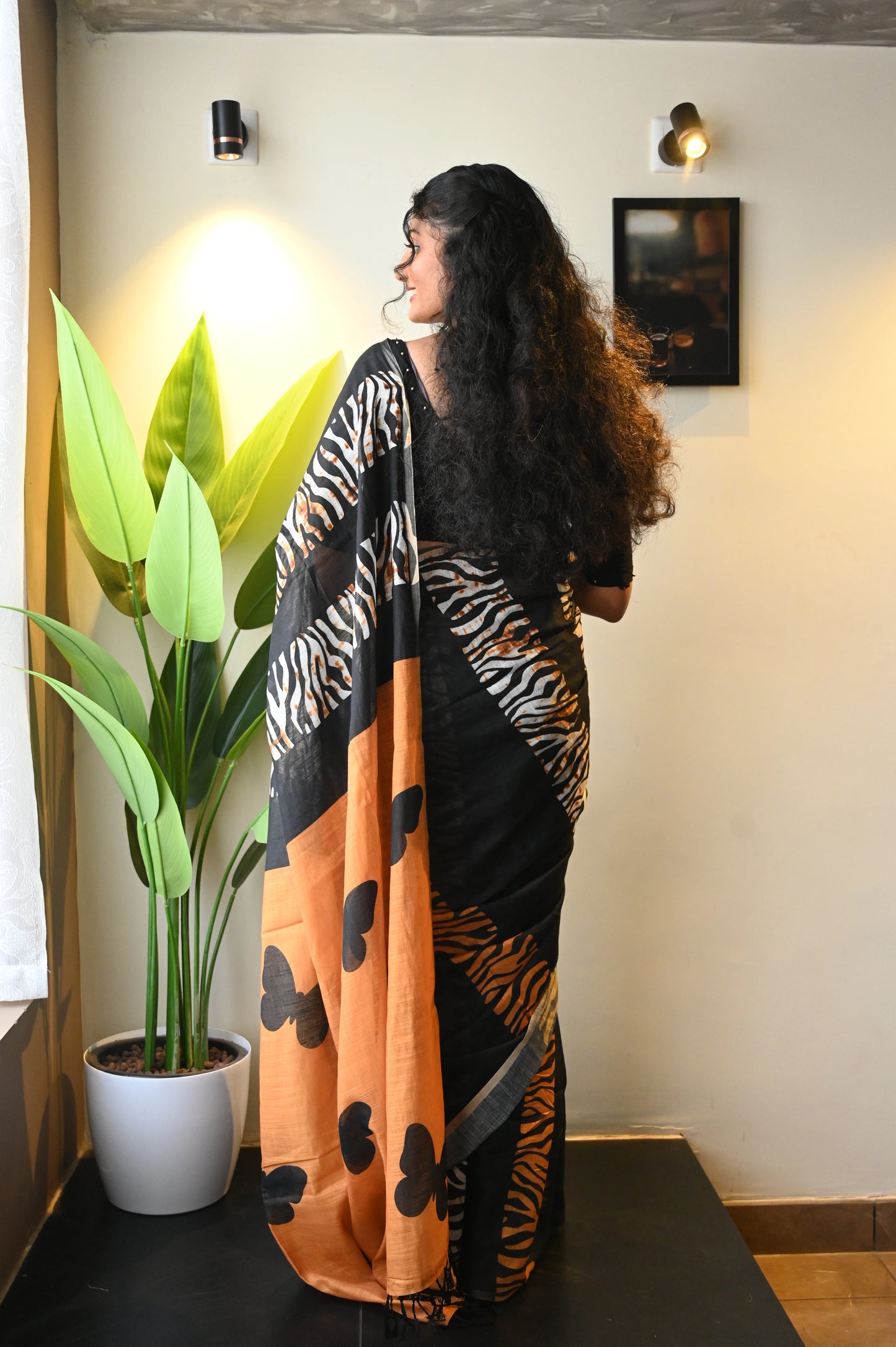 Woman wearing a black and orange patterned saree standing indoors with a plant and wall art in the background.