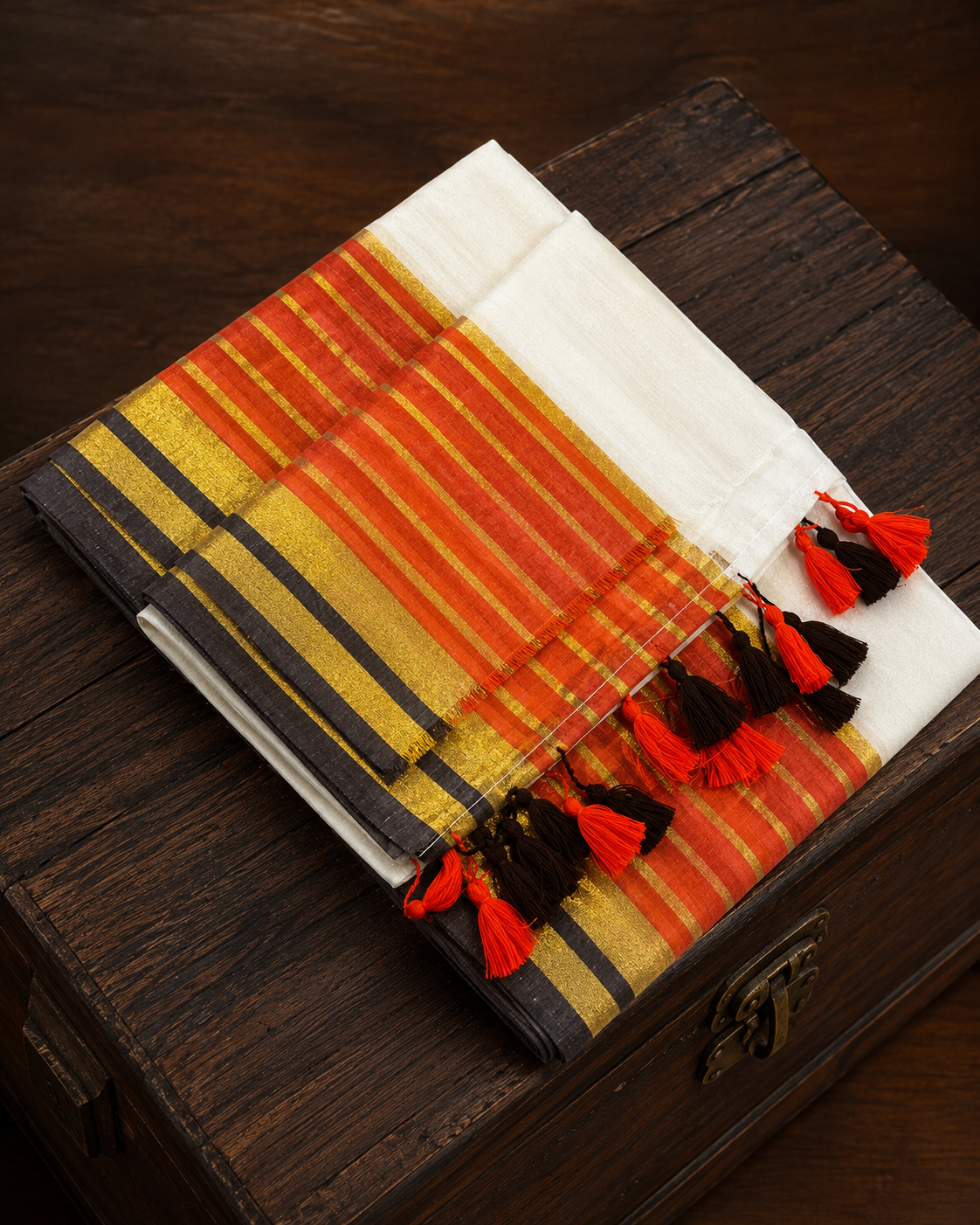A neatly folded traditional white Kerala mulcotton saree with a striking border featuring horizontal stripes of vibrant orange-red, shimmering gold, and deep black, finished with matching orange and black tassels, resting on a dark rustic wooden chest.