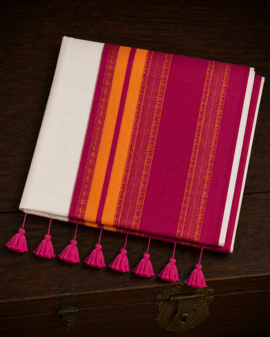 A neatly folded off-white  mul cotton saree featuring a wide, striking border with solid bands of deep magenta and bright orange, accented by intricate orange vertical patterns woven into the magenta weave, and finished with a row of vibrant pink tassels, resting on a rustic dark wooden chest